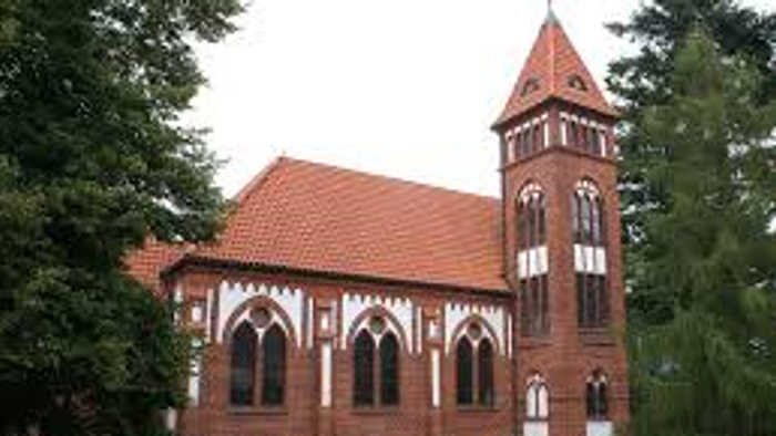 Das Bild zeigt eine rote Backsteinkirche mit hohem Turm und spitzen Fenstern, umgeben von Bäumen und einem Zaun.