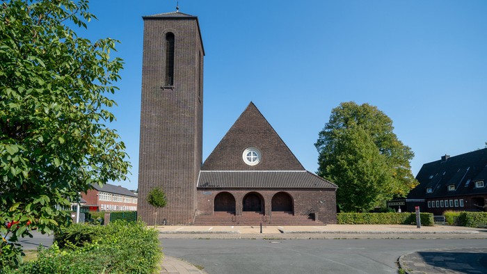 Moderne Backsteinkirche mit hohem Glockenturm und Kreuzsymbol auf dreieckigem Dach, umgeben von Grünflächen.