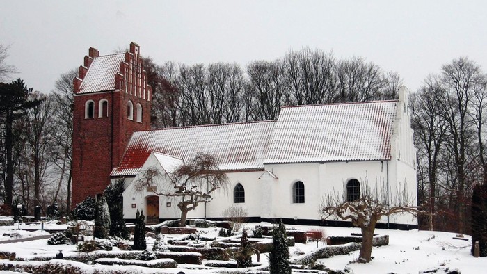 En snedækket kirke med et rødt tårn og et hvidt tag, omgivet af sne og træer