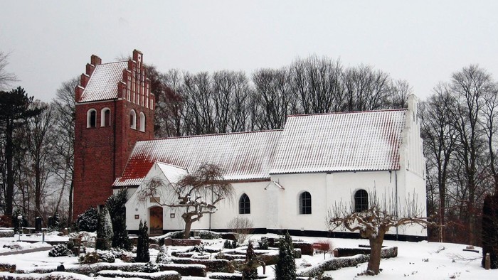 En snedækket kirke med et rødt tårn og et hvidt tag, omgivet af sne og træer