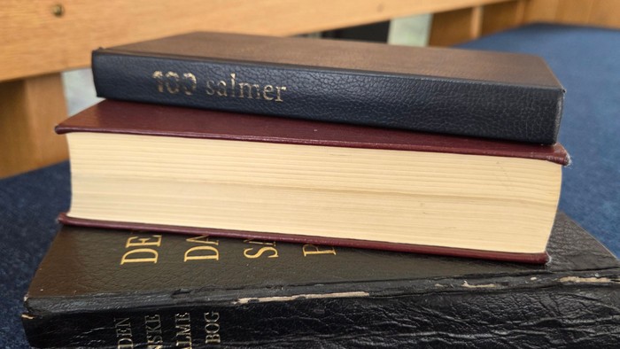 Stak af gamle bøger, herunder en Bibel, en salmebog og en religiøs kommentar, på en blå overflade.