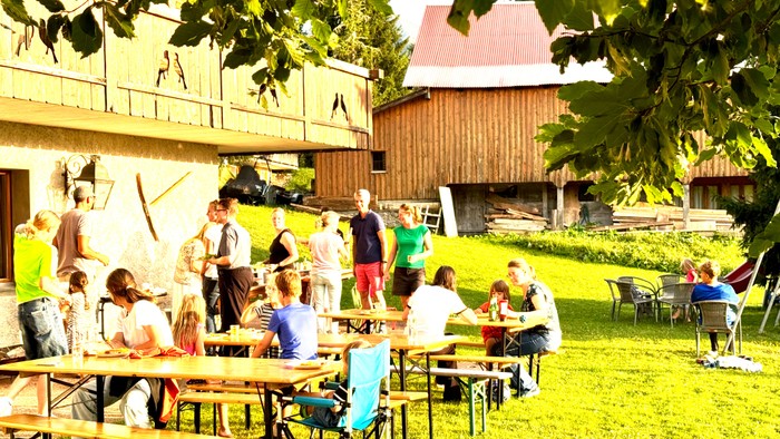 Eine Gruppe von Menschen sitzt an Tischen im Freien vor einem Holzhaus.