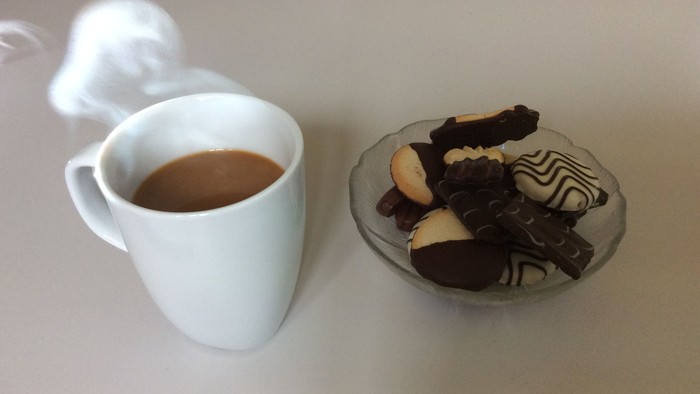 Dampfender Kaffeebecher kombiniert mit einem kleinen Teller mit verschiedenen Pralinen und Gebäck.