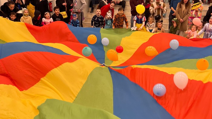 Kinder, die mit einem großen, bunten Fallschirm voller Luftballons bei einer festlichen Indoor-Veranstaltung spielen.
