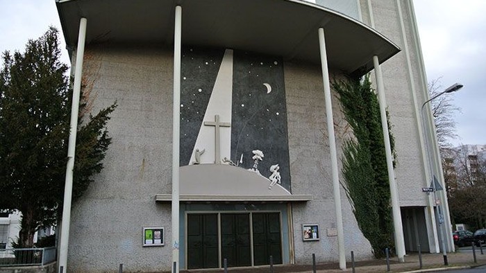 Moderne Kirchenfassade mit einem großen Wandgemälde einer mondbeschienenen Kreuzigungsszene und hohen Stützpfeilern.
