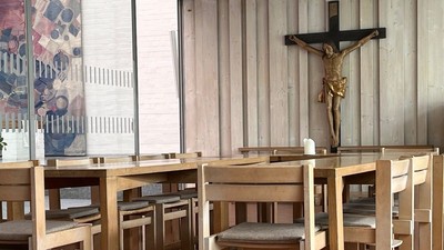 Ein ruhiger Kapelleninnenraum mit hölzernen Kirchenbänken, einem Kruzifix und großen Fenstern, die natürliches Licht hereinlassen.
