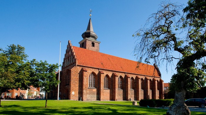 En rød murstenskirke med spir med tårn og grønne træer i forgrunden