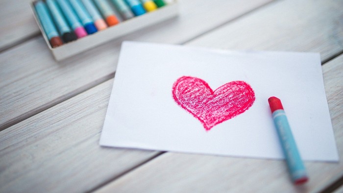 Ein weißes Blatt Papier mit einem pinken Herz, daneben ein blauer Stift und eine Farbstifte-Box auf einem Holztisch