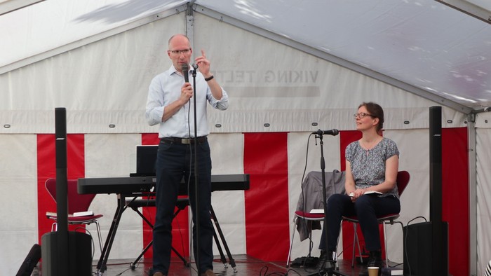 En mand taler i en mikrofon på en scene, mens en siddende kvinde lytter under en udendørsbegivenhed.