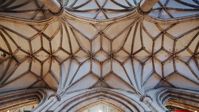 Blick nach oben auf das kunstvolle gotische Gewölbe des Ulmer Münsters. Schlanke steinerne Rippen spannen sich in einem symmetrischen Muster zwischen den Säulen und bilden ein filigranes Netzgewölbe. Dezente florale Bemalungen schmücken die Gewölbeflächen