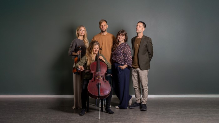 En kvartet af musikere, der står sammen med en cello, en violin og andre instrumenter foran en ensfarvet baggrund.