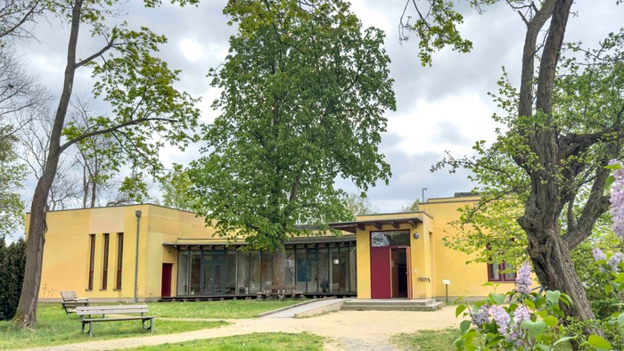 Modernes gelbes Gebäude, umgeben von Grünflächen mit einem Weg und Bänken in einer ruhigen Parkanlage.