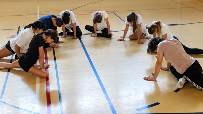 Eine Gruppe von Personen in Sportkleidung, die eine Teamübung oder einen Staffellauf auf einer Indoor-Bahn durchführt.