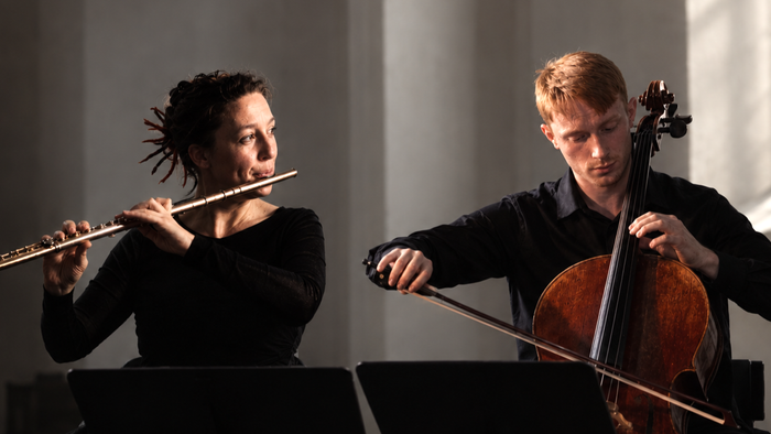 To musikere, der optræder sammen, den ene spiller fløjte og den anden cello i en svagt oplyst koncertscene.