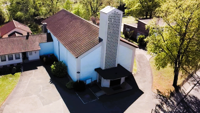 Moderne Kirchenanlage mit hohem, strukturiertem Glockenturm, umgeben von einem gepflasterten Innenhof und Grünflächen.