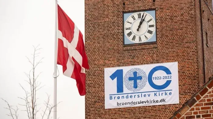Et dansk flag vajer ved siden af en murstensbygning med et ur og et banner med