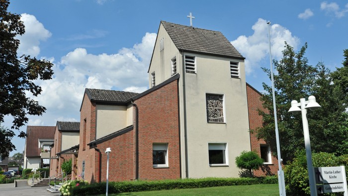 Das Bild zeigt eine Kirche mit einer Fasse aus Backsteinen, teilweise verputzt, mit einem bunten Fenster und einem Kreuz auf dem niedrigen Turm.