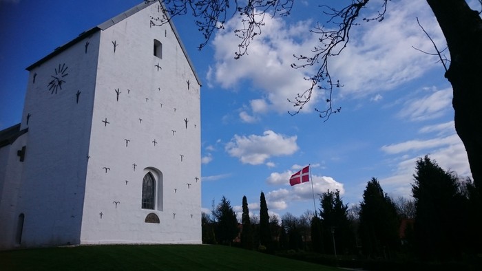 En hvid kirke med dansketragt ved siden af et træ.