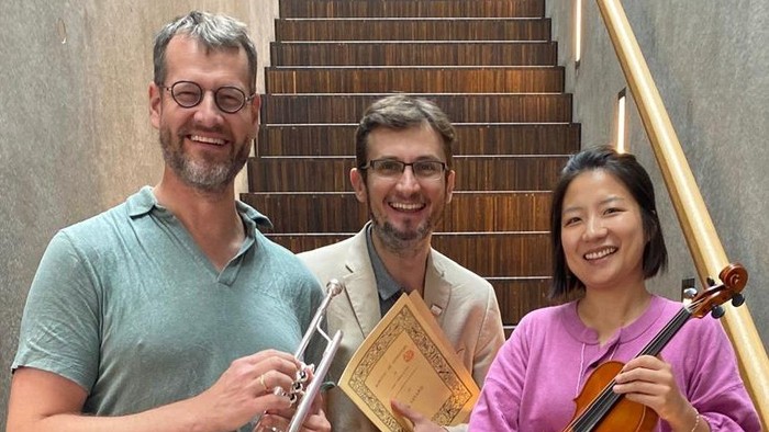 Drei Musiker der Staatskapelle Unter den Linden stehen auf einer Treppe. Eine Asiatin mit ihrer Violine, ein Trompeter mit seinem Instrument und der Pianist mit Noten.