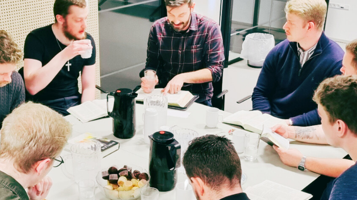En gruppe mænd samlet omkring et bord, hvor de diskuterer med en åben Bibel og snacks til stede.
