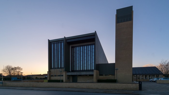 Moderne kirke med højt murstens tårn og stor lodret vinduesfacade i tusmørke.