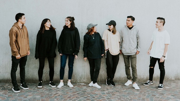 Eine Gruppe von sieben lässig gekleideten Personen, die vor einer schlichten Wand zusammenstehen.