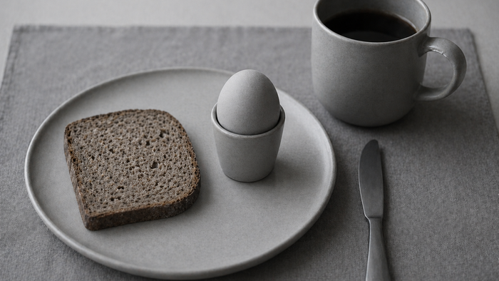 Schlicht gedeckter Frühstücksplatz mit einer Scheibe Brot auf einem Teller, einem Ei im Eierbecher und einer Tasse schwarzem Kaffee daneben; alles in zurückhaltenden Grautönen, ruhig und minimalistisch arrangiert.