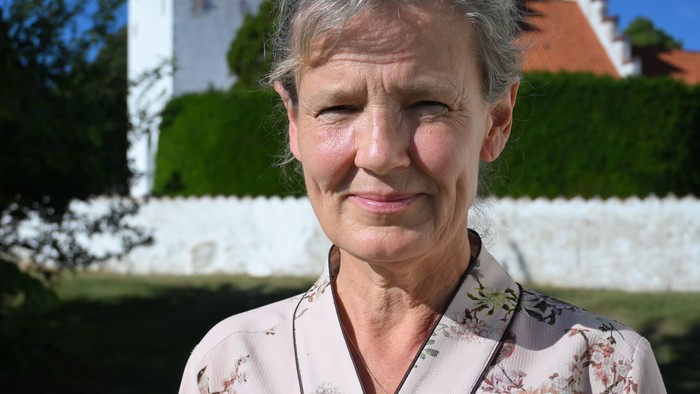 En kvinde i en blomstret cardigan står udendørs nær en hvid kirke under en klar blå himmel.