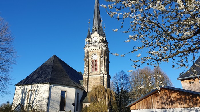 Eine friedvolle Kirche mit hohem Kirchturm steht neben blühenden Bäumen unter einem klaren blauen Himmel.
