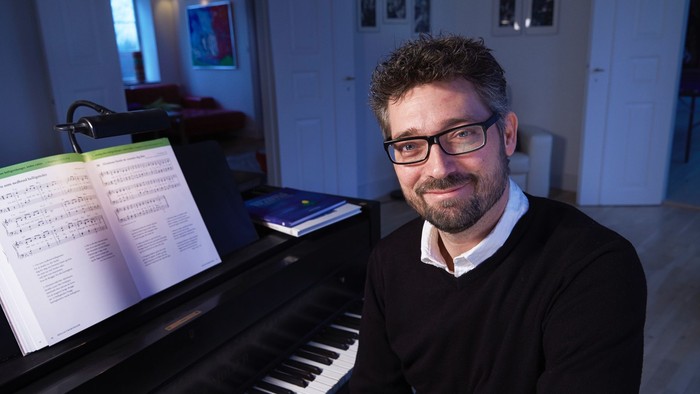 En mand med briller smiler, mens han sidder ved et klaver med nodeark åbent foran sig.