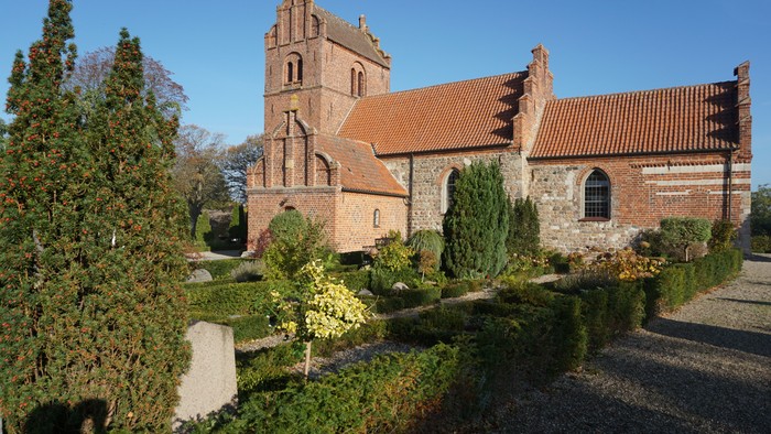 En gammel kirke med teglmur og spiret, omgivet af en have og træer
