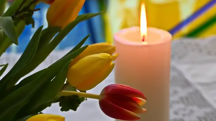 Eine brennende Kerze neben leuchtenden Tulpen auf einer strukturierten Oberfläche mit farbigem Kirchenfensterglas im Hintergrund.