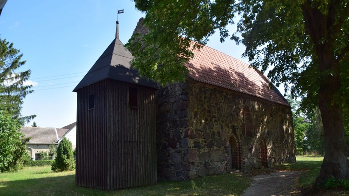 Ein hölzerner Turm und ein Steinbau stehen auf einem grasbewachsenen Grundstück unter Bäumen.