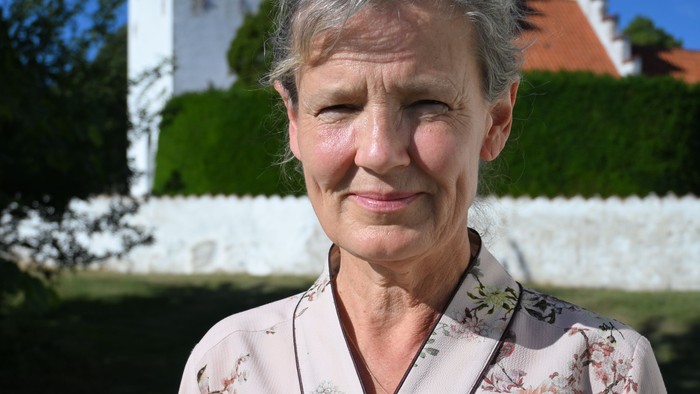 En kvinde i en blomstret cardigan står udendørs nær en hvid kirke under en klar blå himmel.