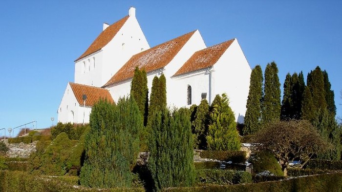 En hyggelig hvid kirke med røde tage gemt mellem nydeligt klippede stedsegrønne buske og et lille vandkunst.