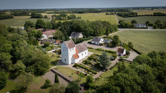 Luftfoto af en idyllisk landsby med en central kirke omgivet af frodig grønne områder og marker.