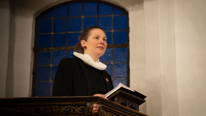 En person i formelt tøj læser op fra en bog ved en prædikestol i en kirke med en baggrund af blå fliser.