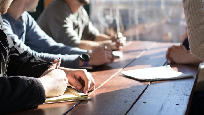 Männer sitzen an einem Tisch und arbeiten mit Notizbüchern und Kaffeetassen.