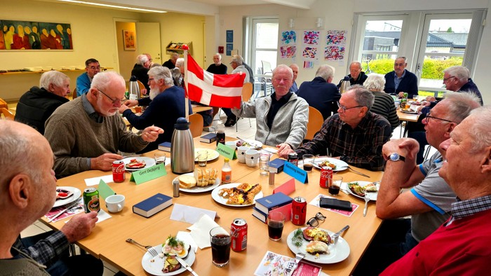 En gruppe ældre mennesker, der nyder et måltid og hygger sig sammen indendørs ved et fællesbord.