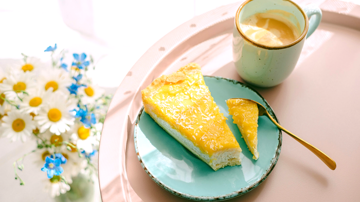 Ein Stück Zitronenkuchen auf einem blauen Teller mit einer Tasse Kaffee und frischen Blumen daneben.