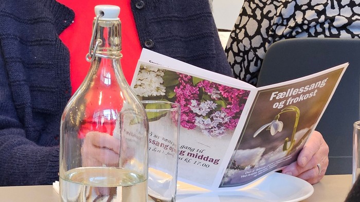 En person holder arrangementsplakater til en sammenkomst med sang og frokost, hvor der står en flaske og et glas i nærheden.