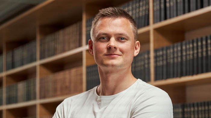 Selvsikker mand poserende med korslagte arme i et bibliotek fyldt med bogreoler.