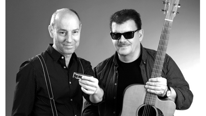 Zwei Männer, einer hält eine Gitarre, der andere ein Handy, beide lächeln.