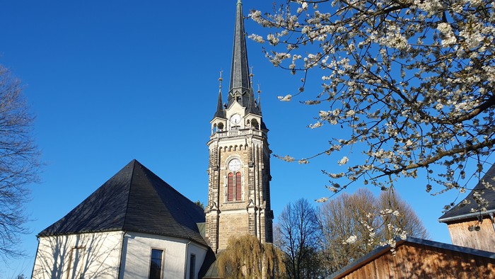 Eine friedvolle Kirche mit hohem Kirchturm steht neben blühenden Bäumen unter einem klaren blauen Himmel.