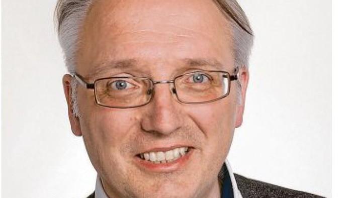 Ein lächelnder Mann mit Brille, der einen Blazer und ein helles Hemd trägt.