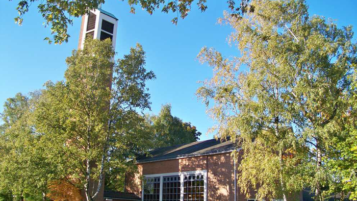 Eine malerische Backsteinkirche mit einem hohen Glockenturm, umgeben von üppigen Herbstbäumen unter einem klaren blauen Himmel.