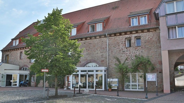 Historisches Steingebäude mit gewölbtem Eingang, umgeben von einem Kopfsteinpflasterhof und Grünflächen.