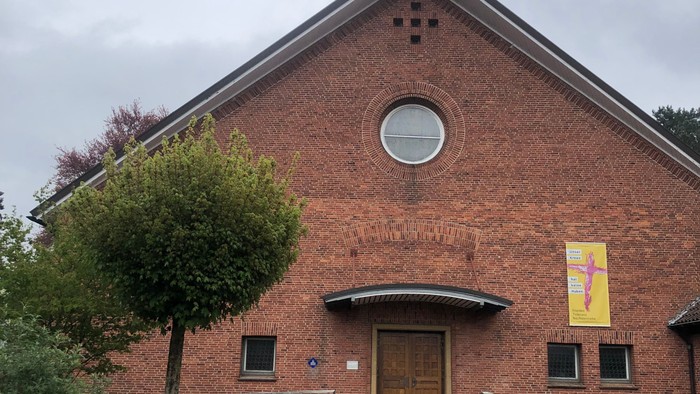 Ziegelsteinkirche mit zentralem Eingang, kleinem Turm und einem bunten Banner über der Tür.