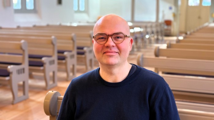 En skaldet mand med briller sidder i en kirkebænk og smiler til kameraet.