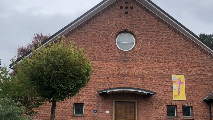 Ziegelsteinkirche mit zentralem Eingang, kleinem Turm und einem bunten Banner über der Tür.